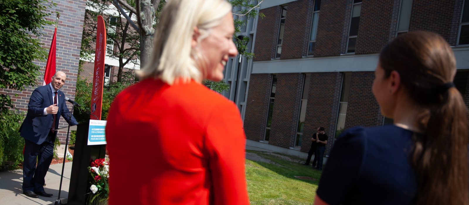 Carleton's President and Vice-Chancellor Benoit-Antoine Bacon stands outdoors at a podium and is smiling at Minister McKenna and Professor Cynthia Cruickshank