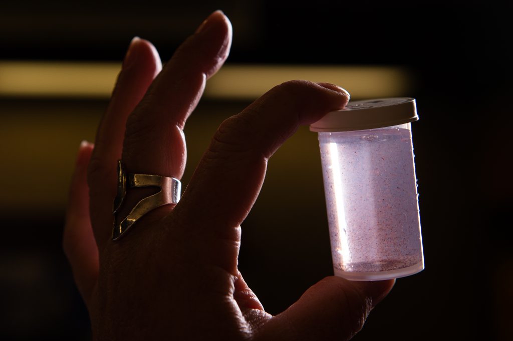 A small clear bottle of pink sand sediment being held to the light between a forefinger and thumb