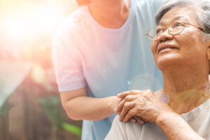 Elder patient setting while holding hand of caretaker who is behind her