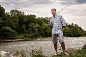 man in plaid shirt standing next to river holding fishing rod over shoulder